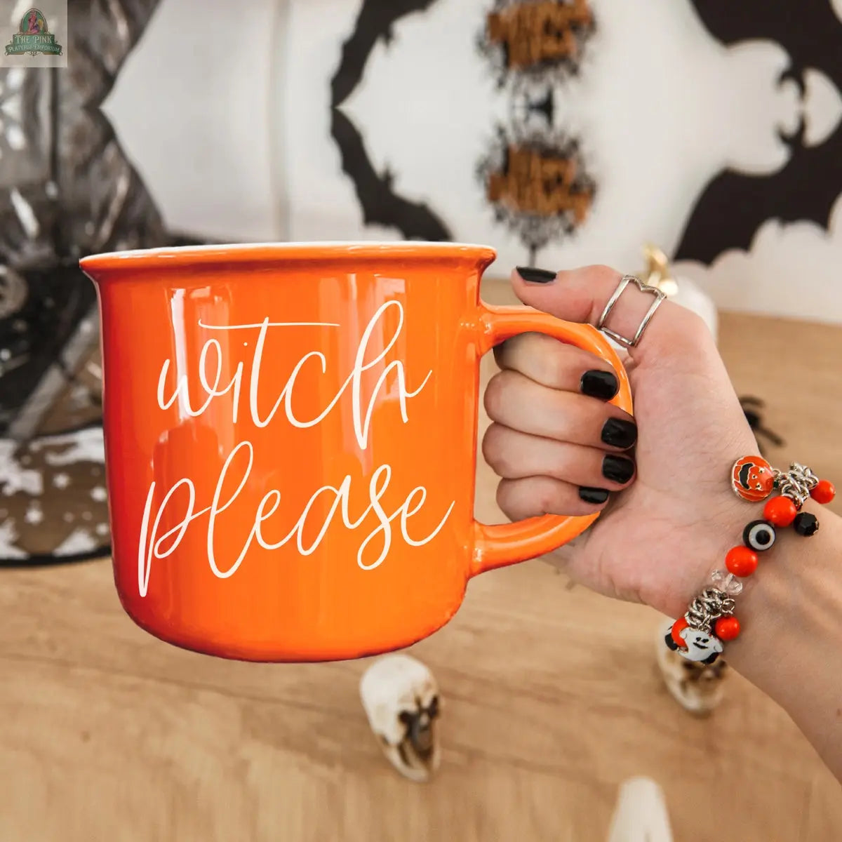 A hand with black nail polish and orange bracelets holds the Witch Please Mug, an orange ceramic mug with "witch please" in white script—perfect for your Halloween coffee moments amid festive decorations.