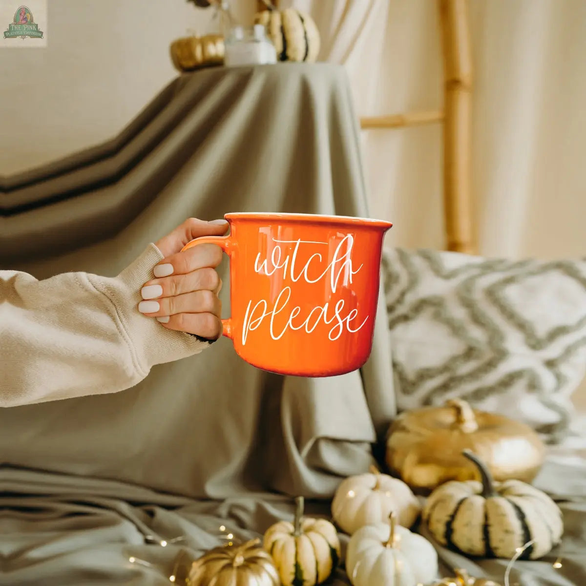 A hand holds the Witch Please Mug, an orange mug featuring the phrase “witch please,” with small white pumpkins and cozy autumn decor in the background.