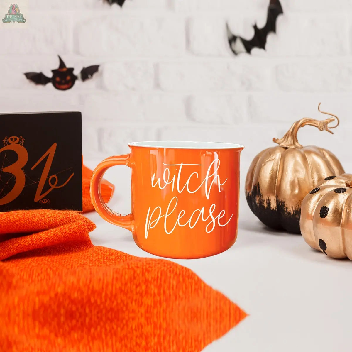 The Witch Please Mug, an orange ceramic cup, rests on an orange cloth beside gold pumpkins, a black 31 sign, and bat décor against a white brick wall—perfect for adding festive Halloween flair to your coffee routine!.