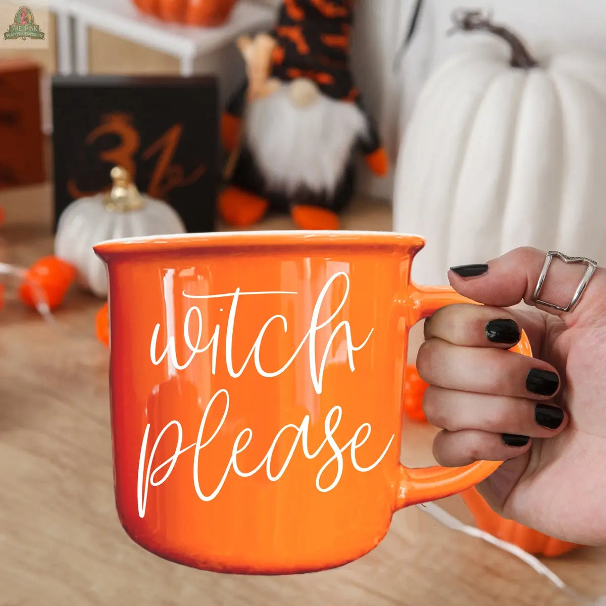 A hand with black nail polish holds the "Witch Please Mug," an orange ceramic mug with "witch please" in white script. Halloween decorations like pumpkins and a gnome are in the background.
