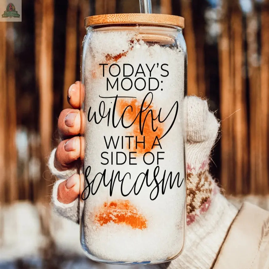 A hand in a knit glove holds the Witchy 20oz tumbler filled with snow, featuring the phrase "Today's mood: witchy with a side of sarcasm." Tall trees blur in the background—perfect as a unique witch gift.
