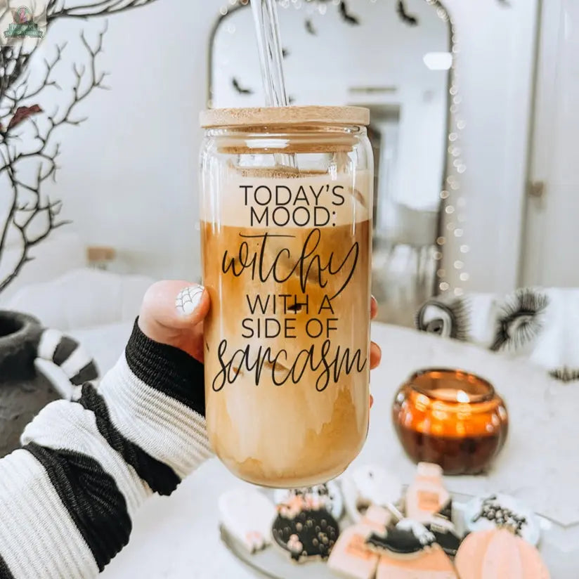 A hand holds the eco-friendly Witchy 20oz glass tumbler filled with iced coffee, featuring “Today’s mood: witchy with a side of sarcasm.” Halloween treats and decor in the background make it a perfect gift for witches.