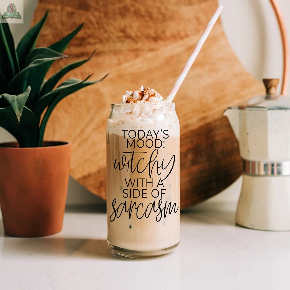 Enjoy iced coffee in the Witchy 20oz glass tumbler, topped with whipped cream and cinnamon, featuring "TODAY’S MOOD: WITCHY WITH A SIDE OF SARCASM." Includes a striped straw, with a plant and coffee pot in the background.