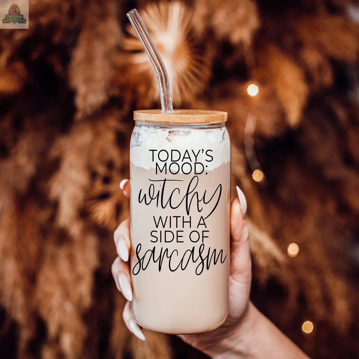 A hand holds the Witchy 20oz—an eco-friendly glass tumbler with bamboo lid and straw, reading "Today's mood: witchy with a side of sarcasm," against blurred warm brown foliage and twinkling lights in the background.