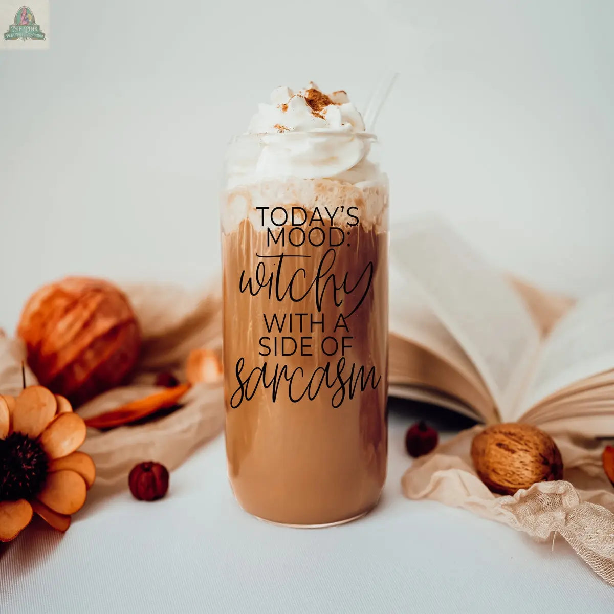 A Witchy 20oz glass tumbler filled with iced coffee, whipped cream, and cinnamon sits on a table with autumn décor. The tumbler reads, “Today’s mood: witchy with a side of sarcasm.” An open book lies in the background.