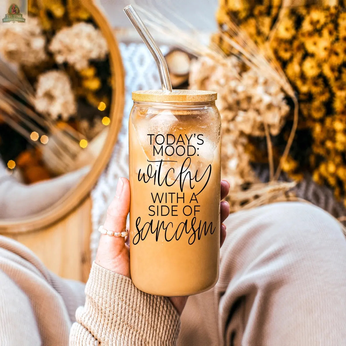 A person in cozy beige clothes holds the Witchy 20oz glass tumbler filled with iced coffee. The label reads, "TODAYS MOOD: WITCHY WITH A SIDE OF SARCASM." Dried flowers and a mirror complete this perfect witchy gift!.