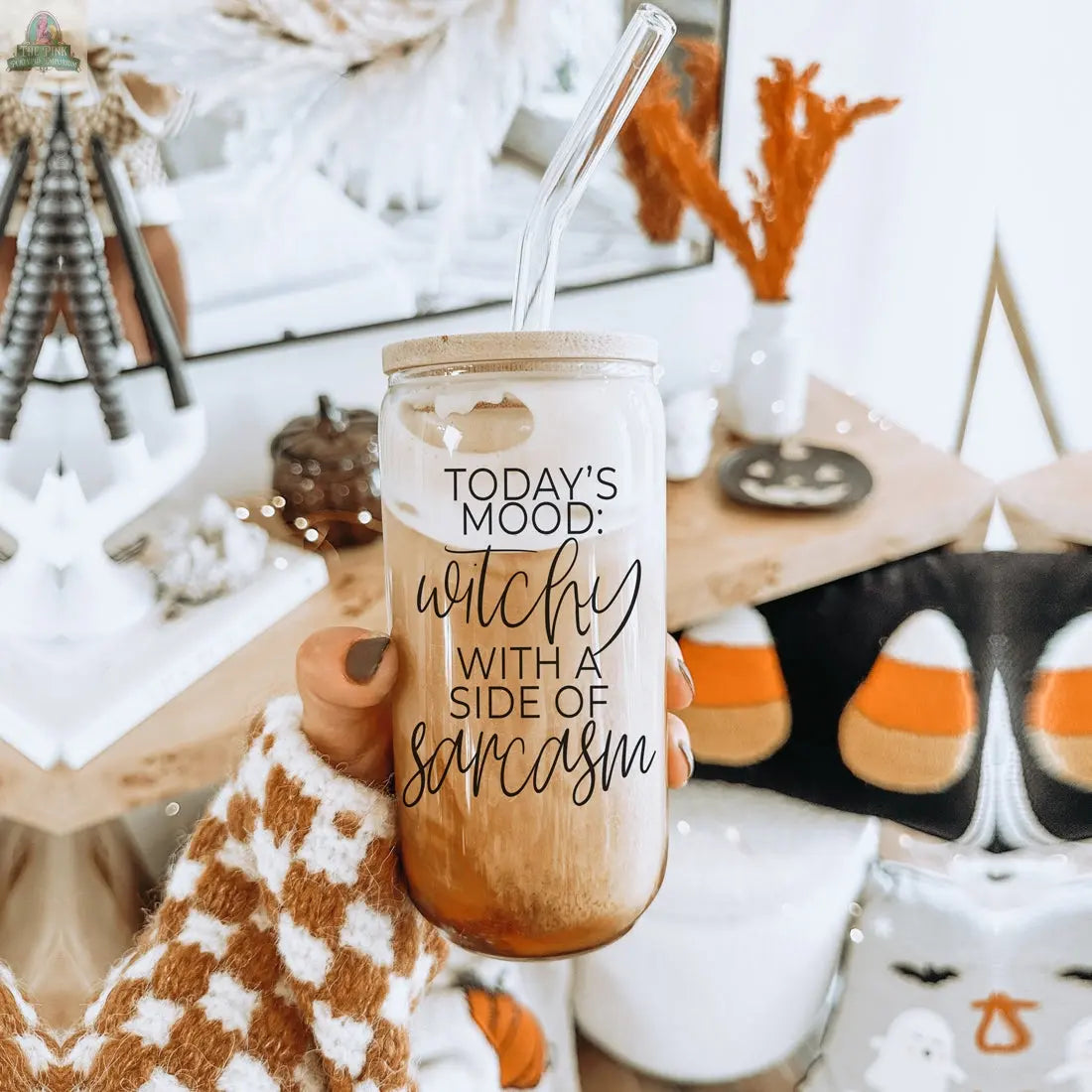 A hand in a brown and white checkered sweater holds the Witchy 20oz glass tumbler, filled with iced coffee. The tumbler reads "Today's mood: witchy with a side of sarcasm." Halloween and autumn decor, like candy corn pillows, complete the scene.