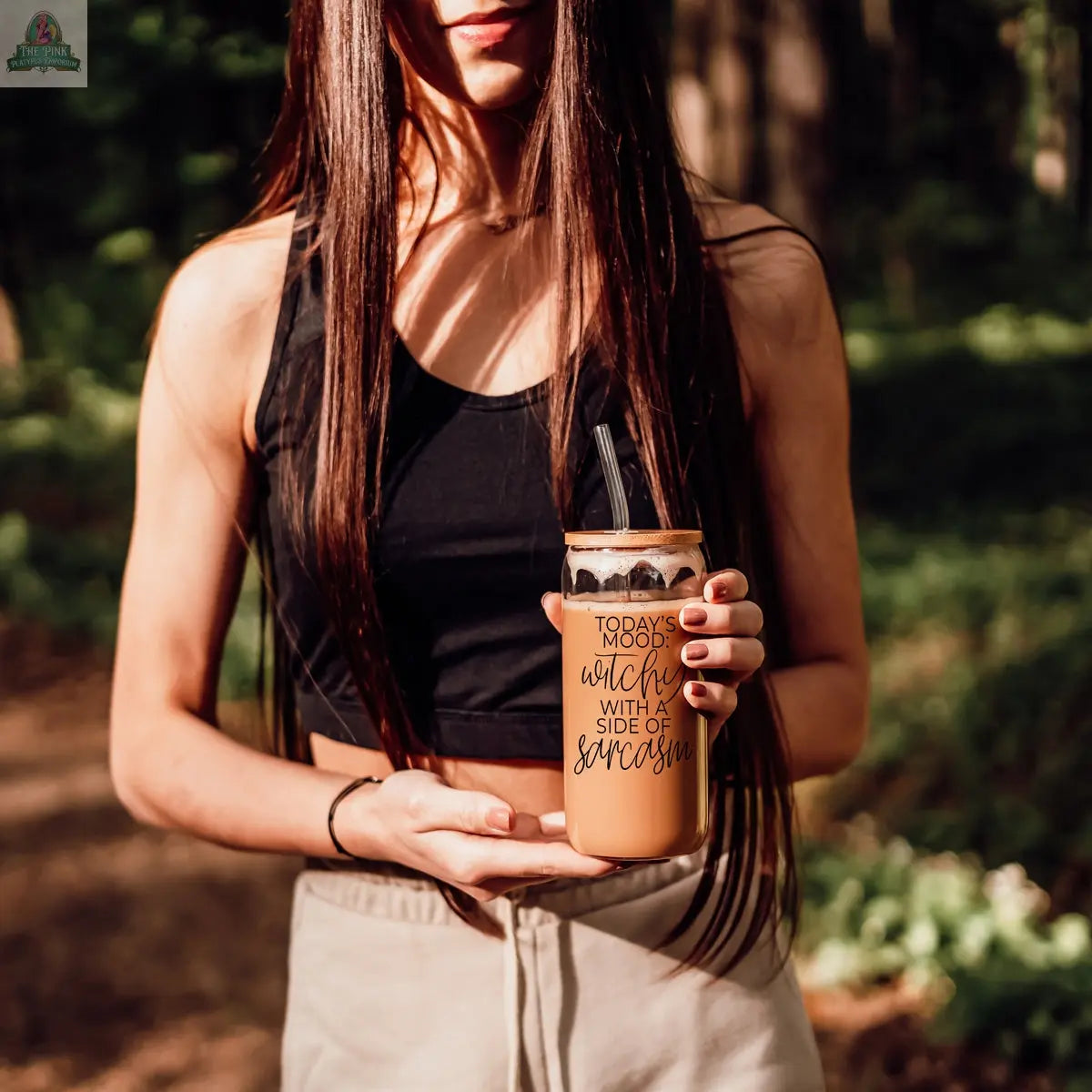 A woman with long brown hair holds a Witchy 20oz glass tumbler that reads "Today's mood: coffee with a side of sarcasm" while standing in a sunlit, wooded outdoor setting.