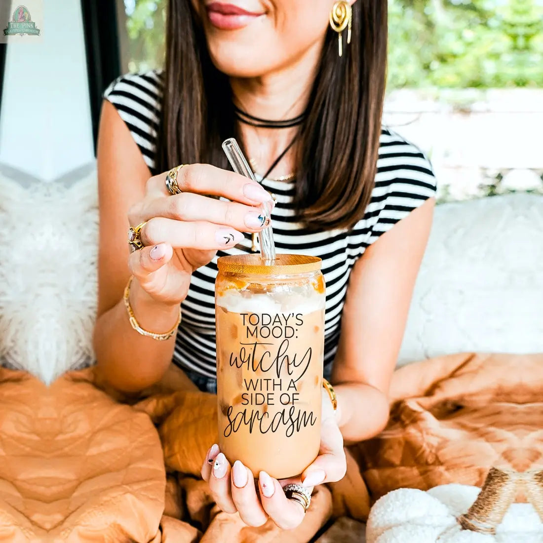 A woman with straight brown hair, wearing a striped shirt and multiple rings, sits on a bed holding the Witchy 20oz tumbler featuring "Today’s mood: witchy with a side of sarcasm"—the perfect witch gift.