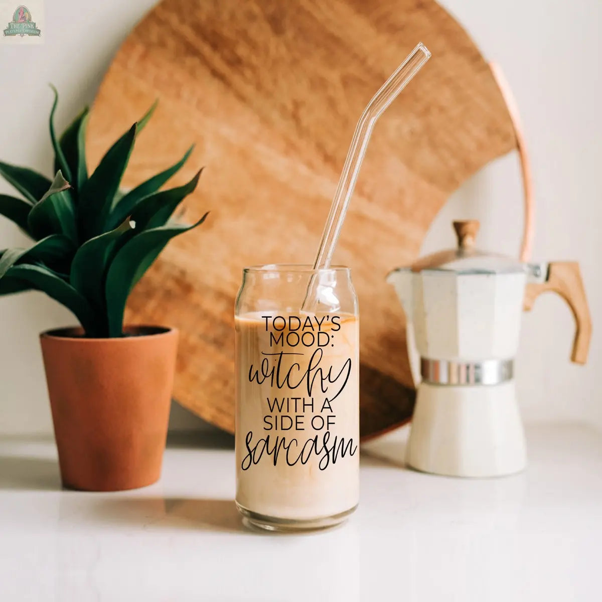 The Witchy 20oz glass tumbler, filled with iced coffee and a glass straw, reads “Today’s mood: witchy with a side of sarcasm.” It’s set on a white surface near a plant, moka pot, and wooden tray.
