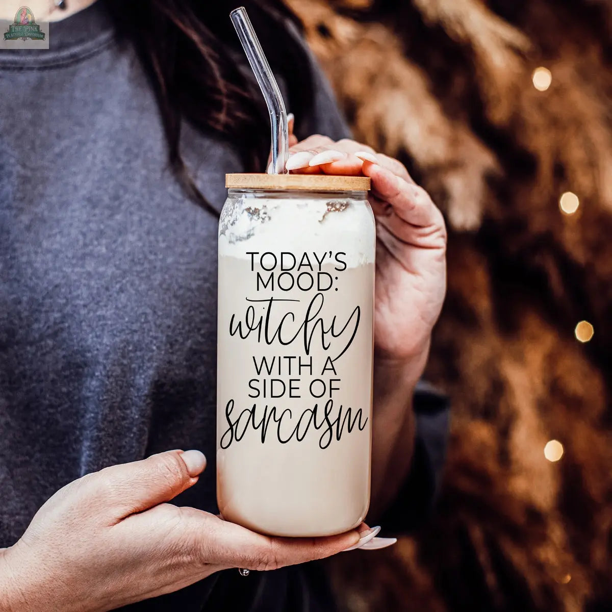 A person holds the Witchy 20oz glass tumbler of iced coffee with a bamboo lid and straw. The glass says, "Today's mood: witchy with a side of sarcasm." Warm brown tones and soft lights blur behind—an ideal eco-friendly witch gift.