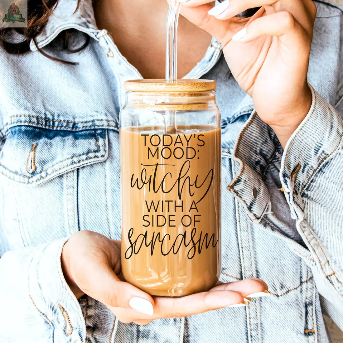 A person in a denim jacket holds the Witchy 20oz eco-friendly tumbler, featuring a bamboo lid and straw, filled with iced coffee. The glass reads: Today's mood: witchy with a side of sarcasm.