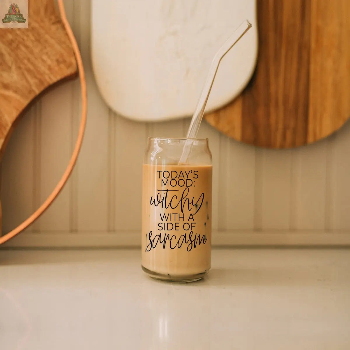 A glass of iced coffee with a clear straw sits on a counter in a Witchy 20oz tumbler labeled "Today's mood: witchy with a side of sarcasm." Wooden and white cutting boards hang on the wall in the background.