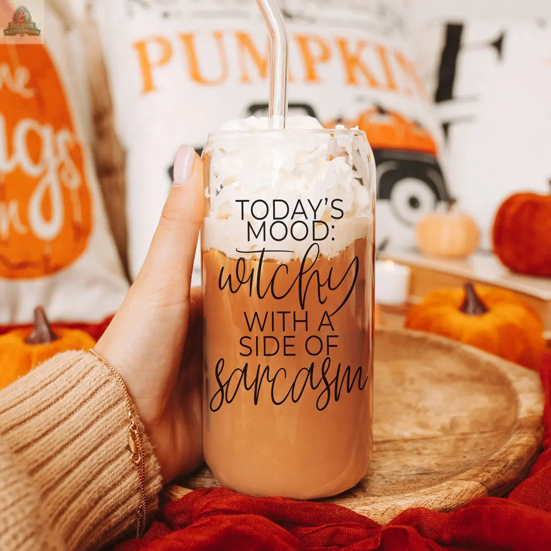 A hand holds the Witchy 20oz glass tumbler filled with iced coffee, whipped cream, and a straw. The tumbler features “Today's mood: witchy with a side of sarcasm.” Fall decor and pumpkins make it the perfect witch gift scene.