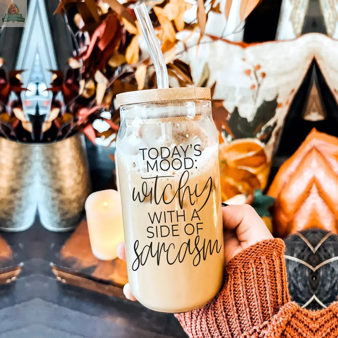 A hand in an orange sweater holds the Witchy 20oz glass tumbler, filled with iced coffee and a straw. The jar reads "Today's mood: witchy with a side of sarcasm." Autumn leaves and a lit candle decorate the background.