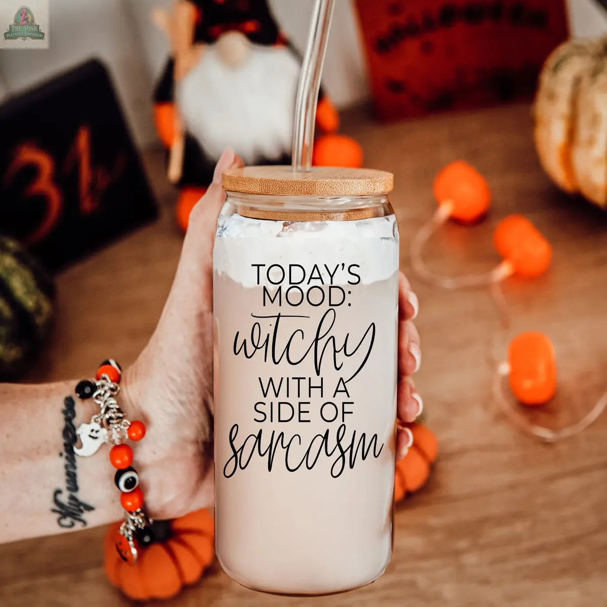 A hand holds the Witchy 20oz glass tumbler with bamboo lid and straw, filled with a light drink. The tumbler reads, "Today’s mood: witchy with a side of sarcasm." Halloween décor and mini pumpkins are in the background.