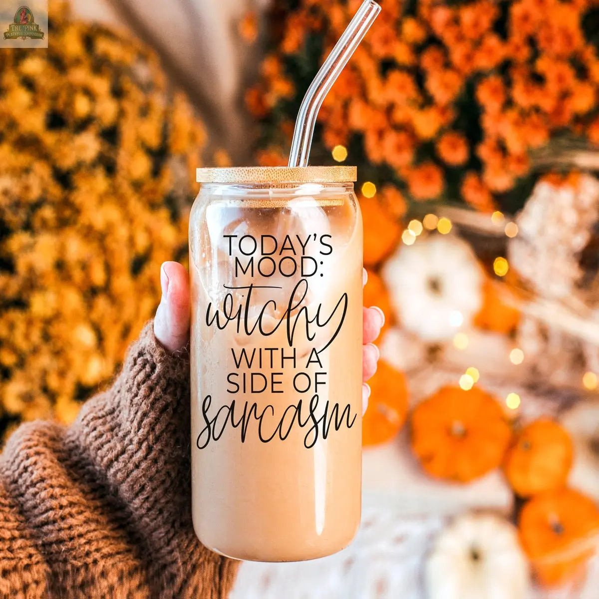 A hand in a cozy brown sweater holds the Witchy 20oz tumbler of iced coffee with a metal straw, featuring the phrase "Today's mood: witchy with a side of sarcasm." Orange flowers and pumpkins blur in the background.
