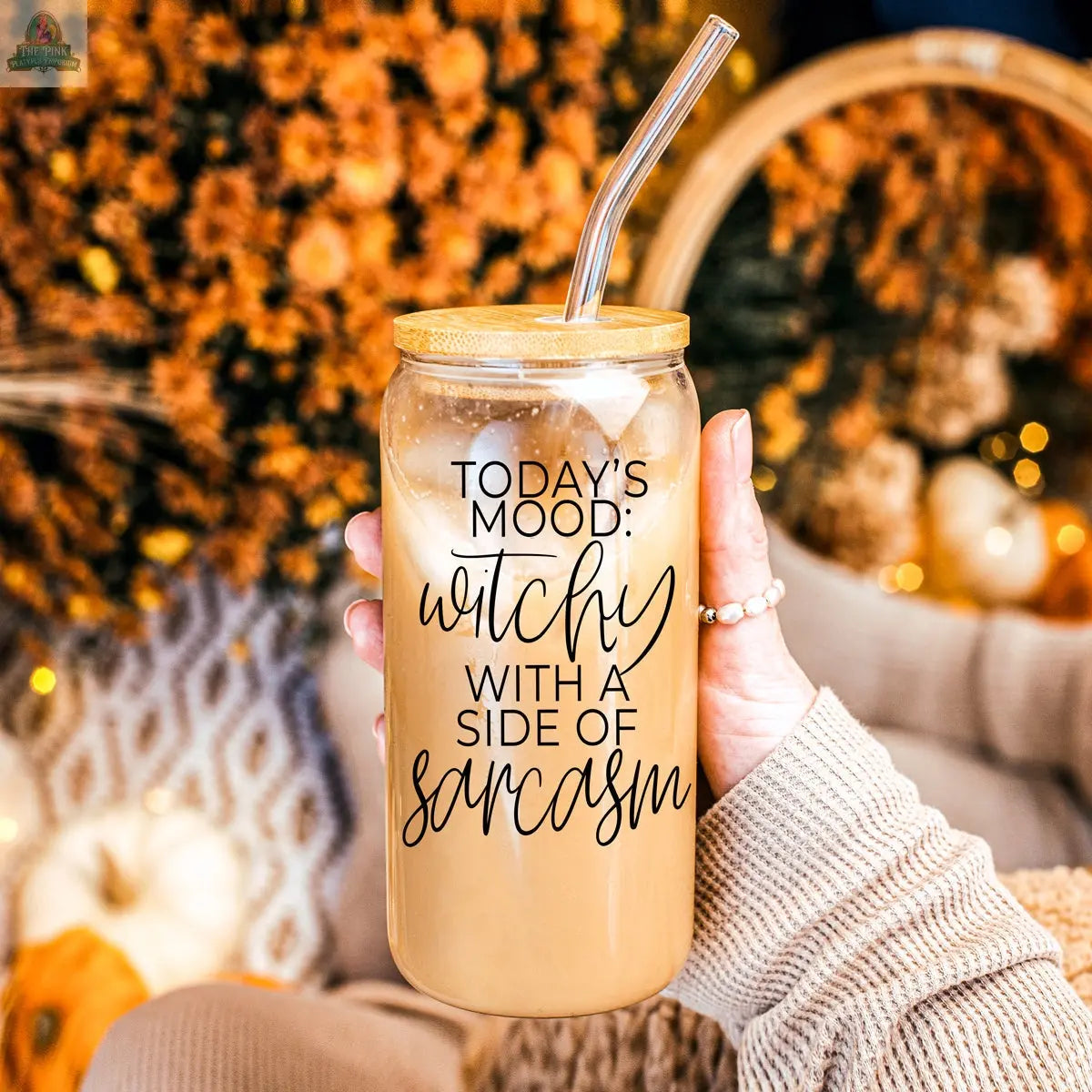 A hand holds the Witchy 20oz glass tumbler filled with iced coffee and a metal straw. The glass says, “Todays mood: witchy with a side of sarcasm,” with blurred autumn décor—orange flowers and pumpkins—in the background.