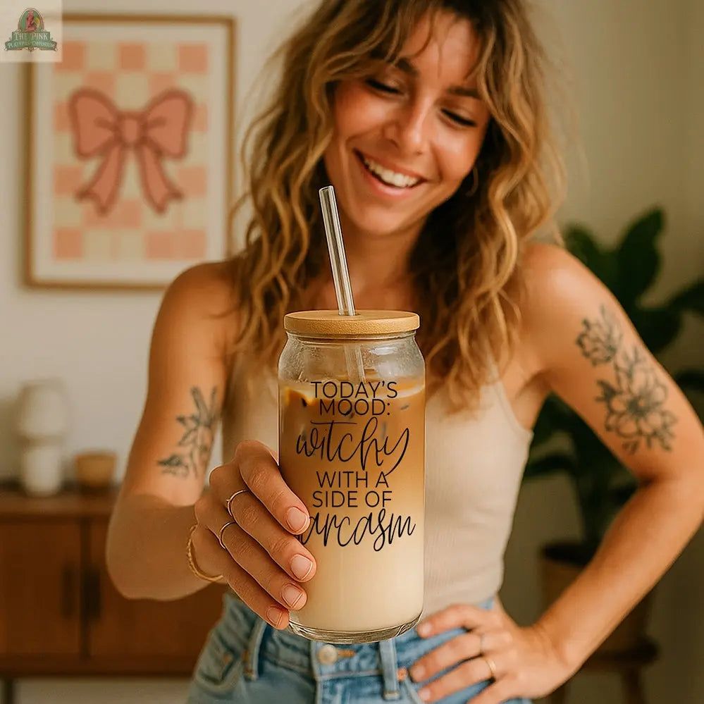 A smiling woman with wavy hair holds the Witchy 20oz eco-friendly tumbler filled with iced coffee. The glass says, "Todays mood: catchy with a side of sarcasm." She stands in a cozy room—an ideal witch gift for playful spirits.