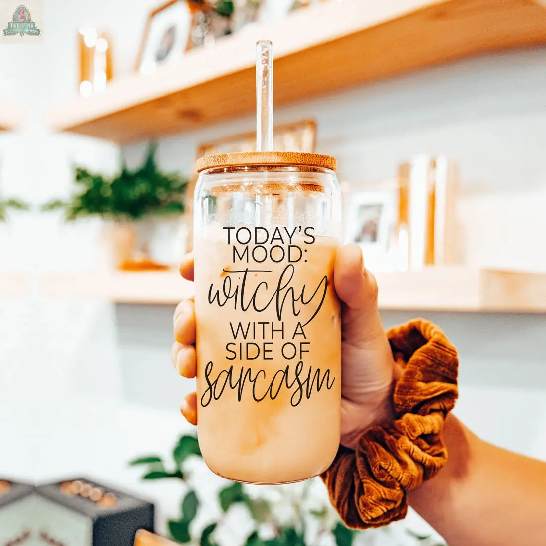 A hand holds the Witchy 20oz glass tumbler filled with iced coffee and a straw. The jar says "Today's mood: witchy with a side of sarcasm." A brown scrunchie is on the wrist, with shelves of plants and decor in the background.