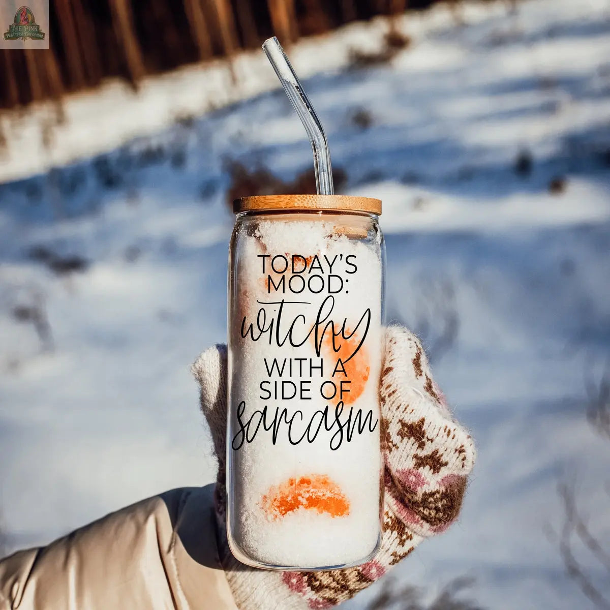 A gloved hand holds the Witchy 20oz glass tumbler, topped with a bamboo lid and straw and filled with a snowy drink. The tumbler says "Today’s mood: witchy with a side of sarcasm." Snowy trees blur in the background—a great witch gift.