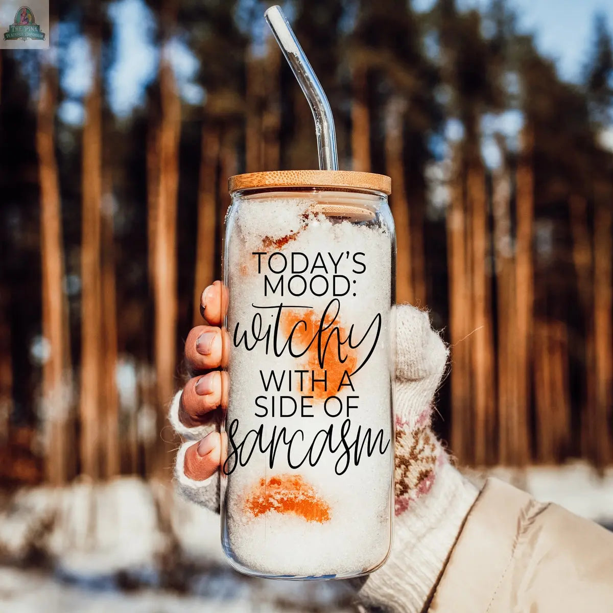 A gloved hand holds the Witchy 20oz eco-friendly tumbler with a bamboo lid and straw, filled with a snowy drink. The glass reads, "Today's mood: witchy with a side of sarcasm," against blurred pine trees and snow in the background.