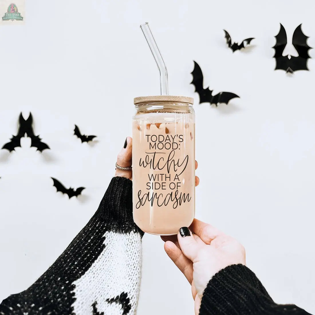 A hand holds a Witchy 20oz glass tumbler filled with iced coffee and a straw. The jar says, “Today’s mood: witchy with a side of sarcasm.” Black bat decor is on a white wall, and the hand wears a black-and-white sweater.