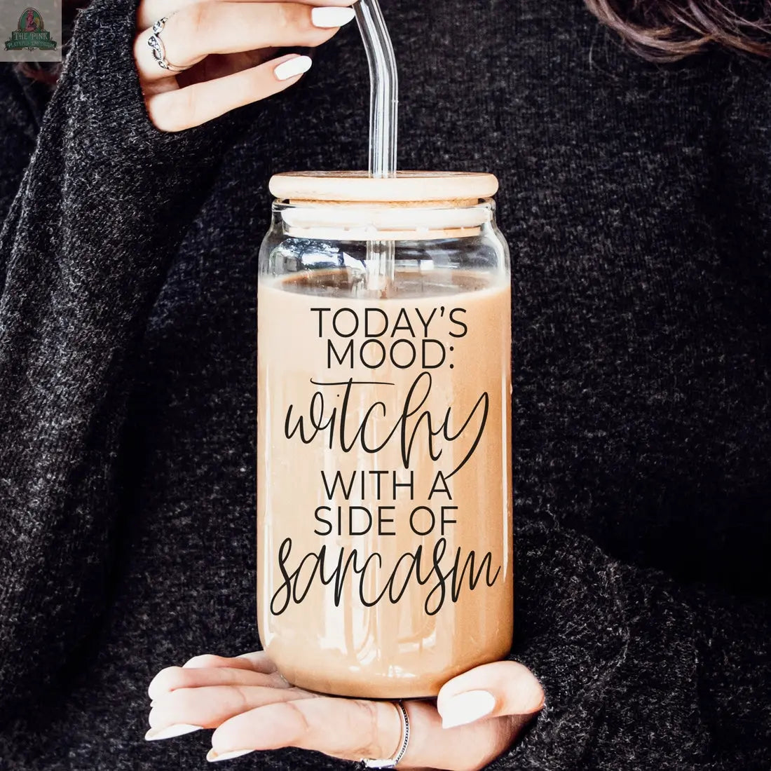 A person in a dark sweater holds the Witchy 20oz eco-friendly glass tumbler of iced coffee with a straw. The tumbler reads, “Today’s mood: witchy with a side of sarcasm”—perfect as a unique witch gift.