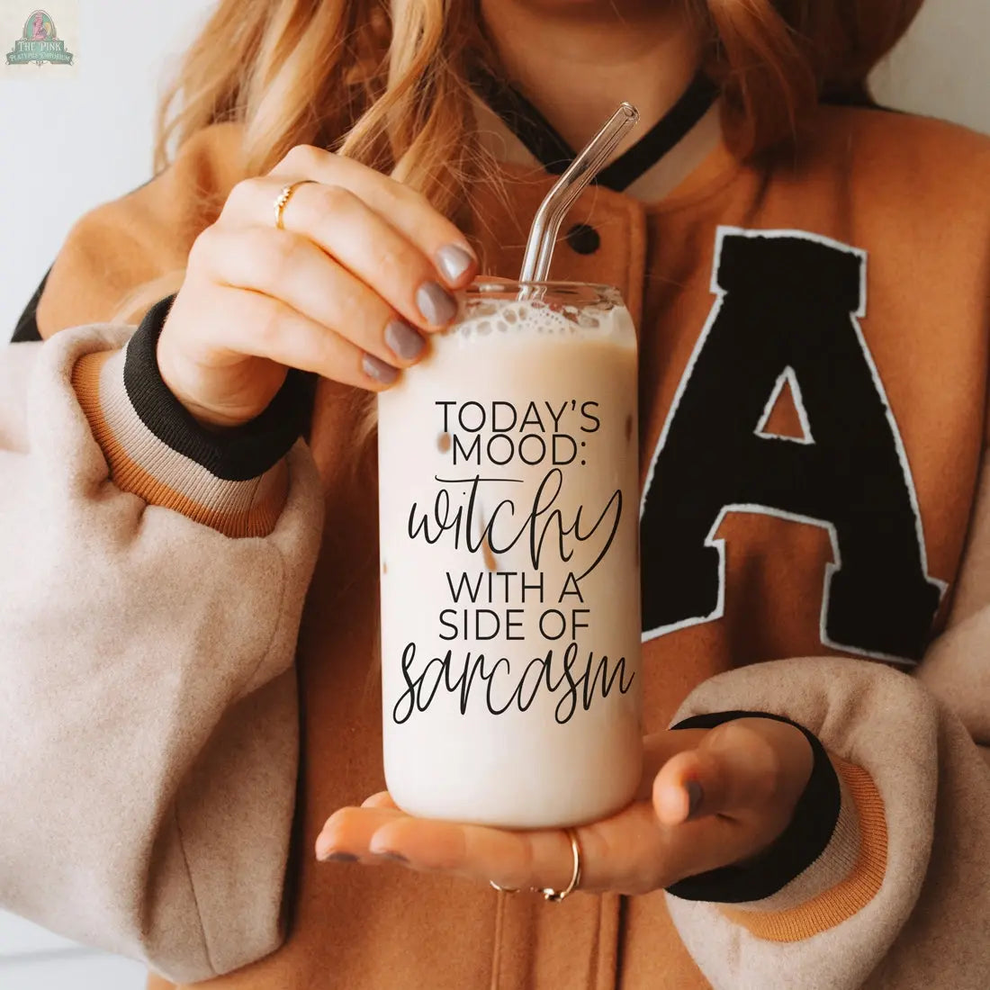 A woman holds the Witchy 20oz glass tumbler with iced coffee and a glass straw. The tumbler says "TODAYS MOOD: witchy WITH A SIDE OF sarcasm"—a unique, eco-friendly gift. She wears a tan and black varsity jacket with an A.