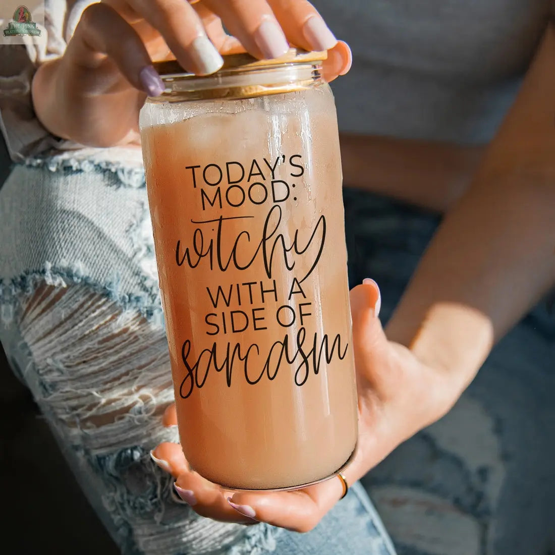 A person in ripped jeans holds the Witchy 20oz glass tumbler filled with a light brown drink. The tumbler displays "TODAYS MOOD: witchy WITH A SIDE OF sarcasm.