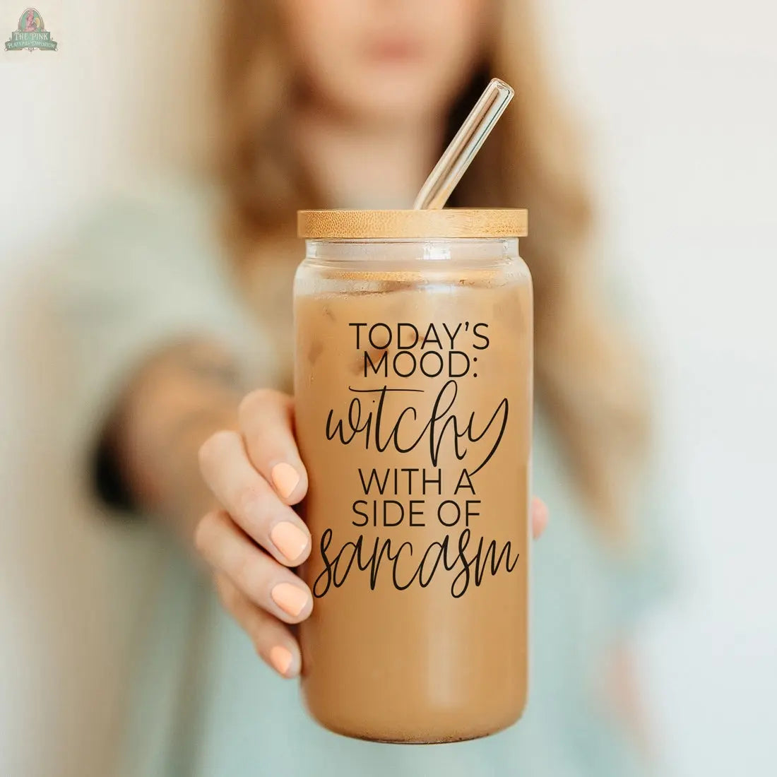 A person holds the Witchy 20oz eco-friendly tumbler filled with iced coffee. The glass jar with bamboo lid and straw reads, "Today's mood: witchy with a side of sarcasm"—a perfect witch gift. A softly blurred background adds charm.