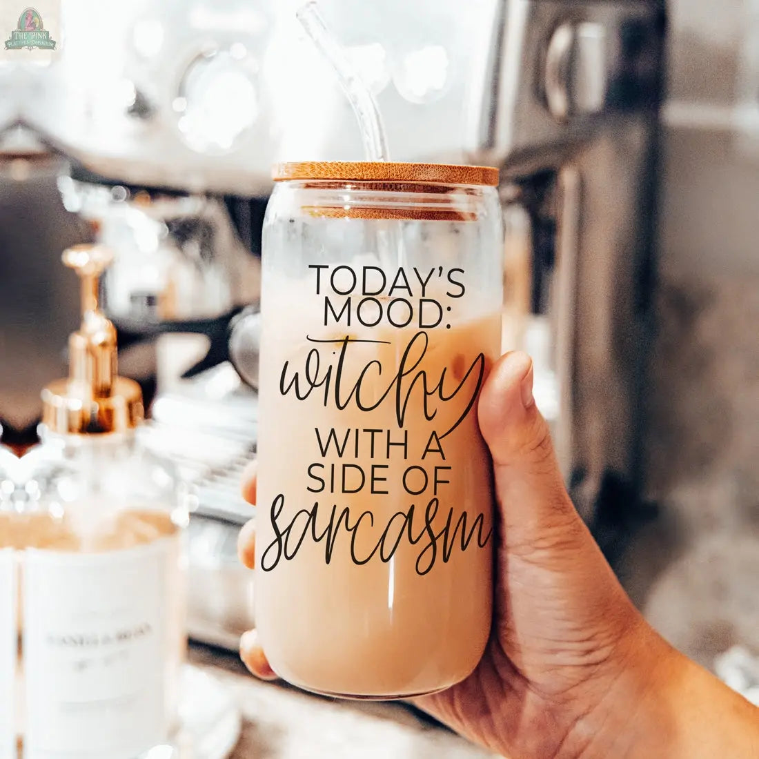 A hand holds the Witchy 20oz glass tumbler with bamboo lid and straw, filled with iced coffee. The tumbler reads: "Today's mood: witchy with a side of sarcasm." A coffee machine and syrup bottle are in the background.