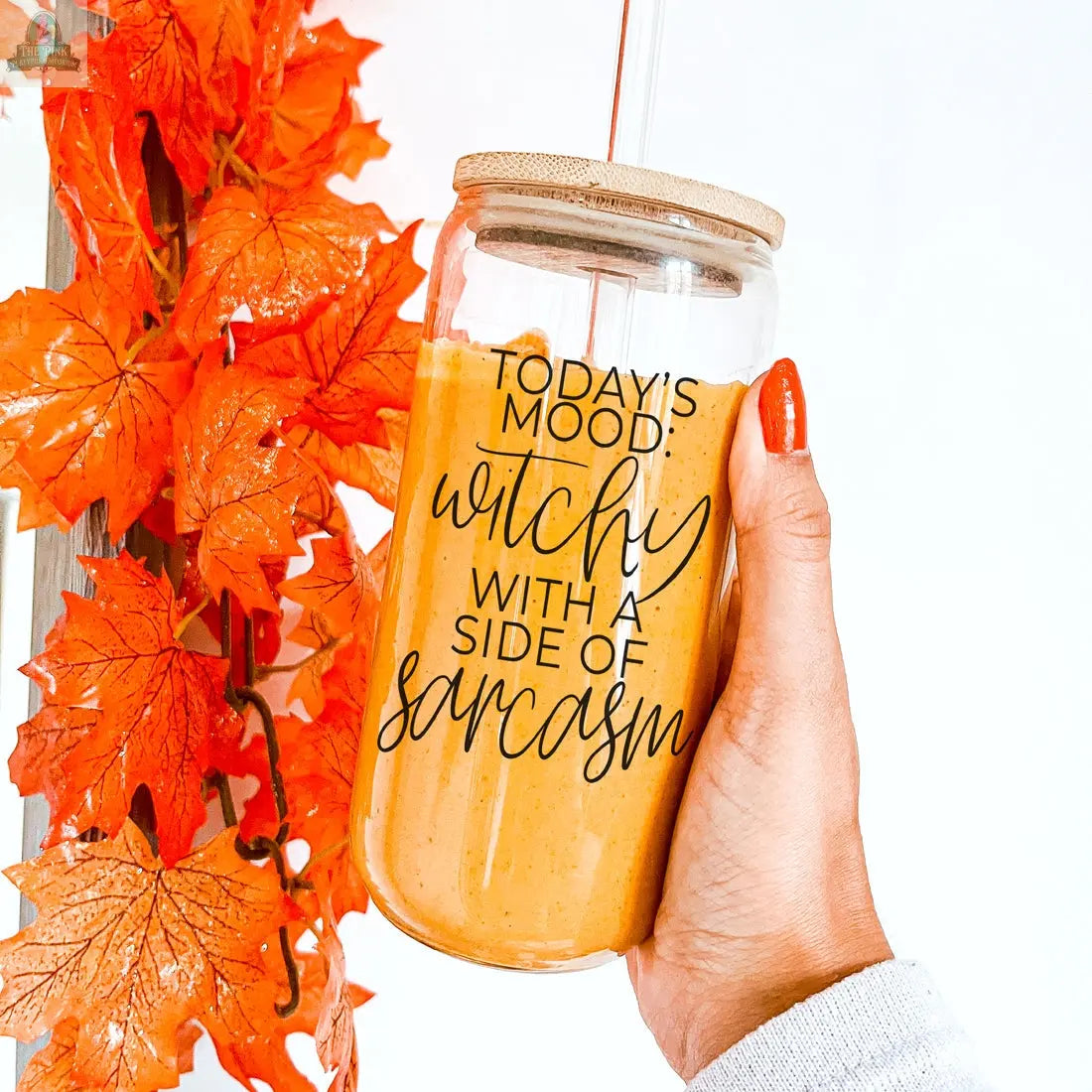 A hand with orange-painted nails holds the Witchy 20oz tumbler, featuring a bamboo lid and straw, filled with an orange drink. It reads “TODAYS MOOD: witchy with a side of sarcasm.” Bright orange fall leaves are beside it.