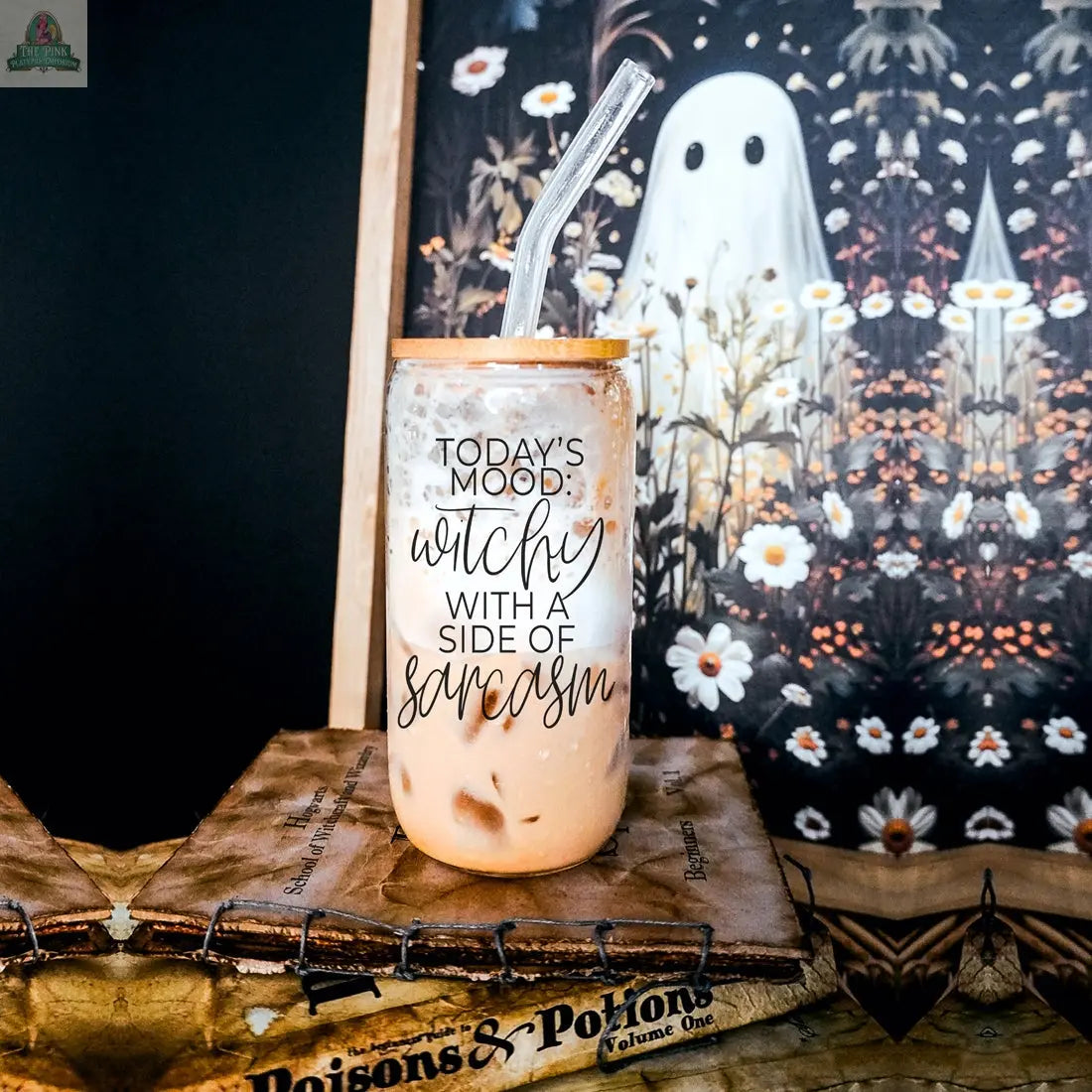 The Witchy 20oz glass tumbler with clear straw sits atop vintage books labeled Poisons & Potions. Its eco-friendly design says, "Today's mood: witchy with a side of sarcasm." Spooky ghost and wildflower art decorates the backdrop.