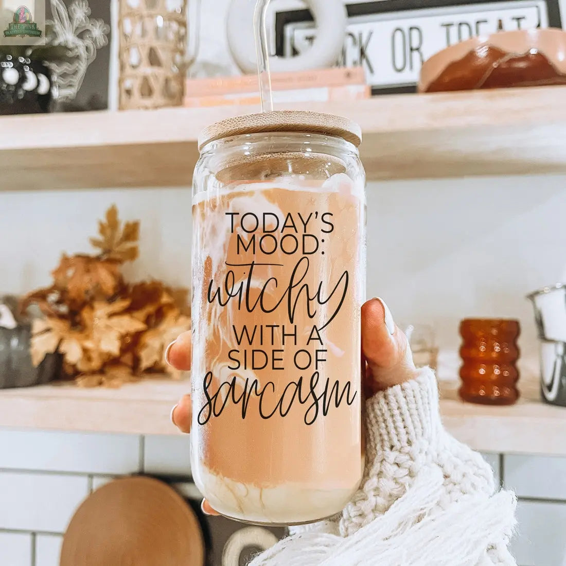 A hand holds the Witchy 20oz glass tumbler filled with a creamy drink and straw. The tumbler features the phrase "Today's mood: witchy with a side of sarcasm" and is set against a background of autumn decor and kitchen items.