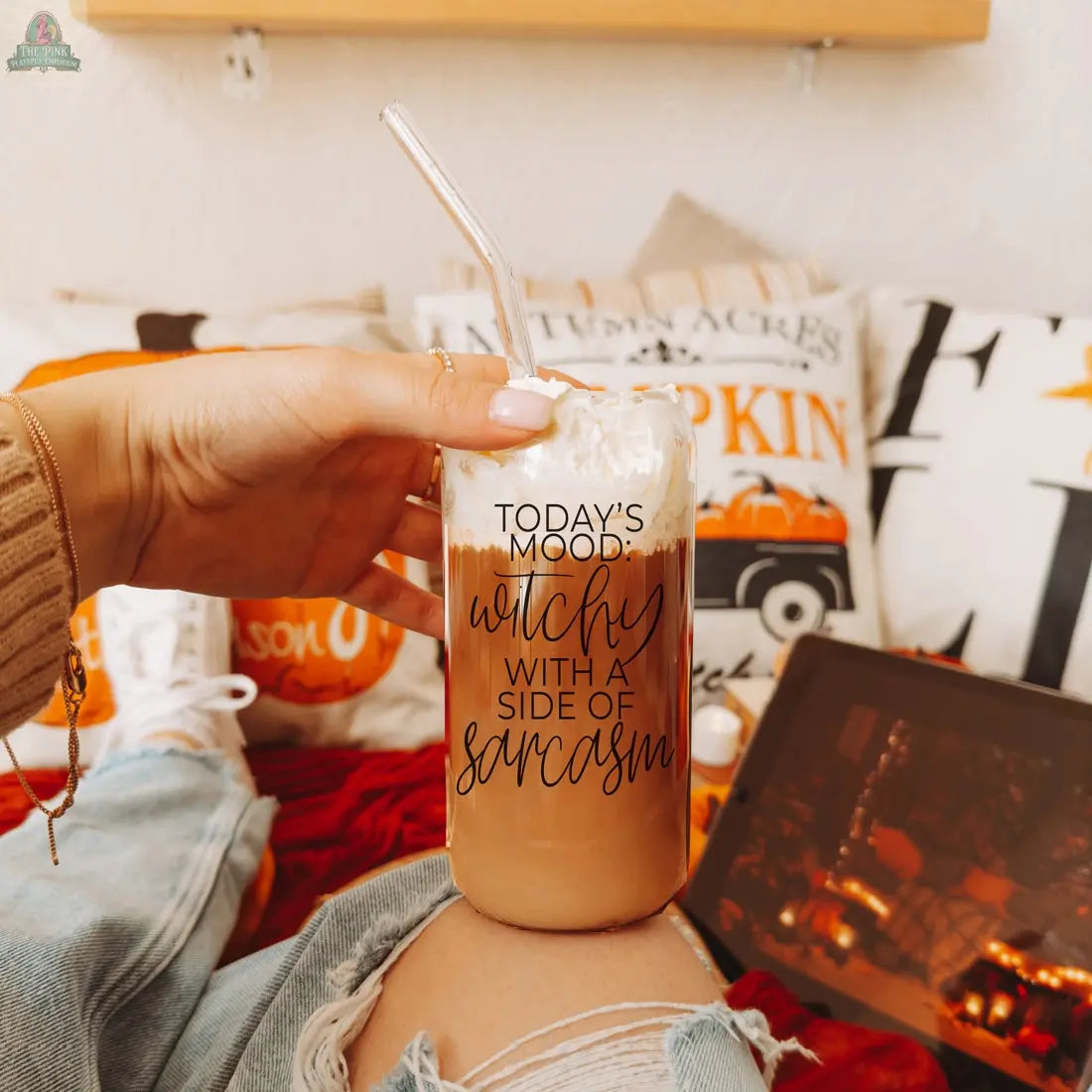 A hand holds a Witchy 20oz glass tumbler filled with iced coffee, topped with whipped cream and a clear straw. The glass says "TODAYS MOOD: witchy with a side of sarcasm." Cozy autumn decor and a fireplace are in the background.