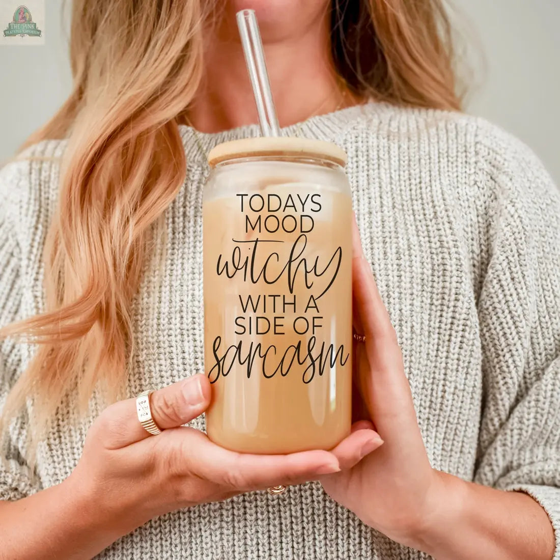 A woman in a gray knit sweater holds the Witchy 20oz glass tumbler with a straw, featuring the phrase "TODAYS MOOD WITCHY WITH A SIDE OF SARCASM," and a light-colored drink inside.