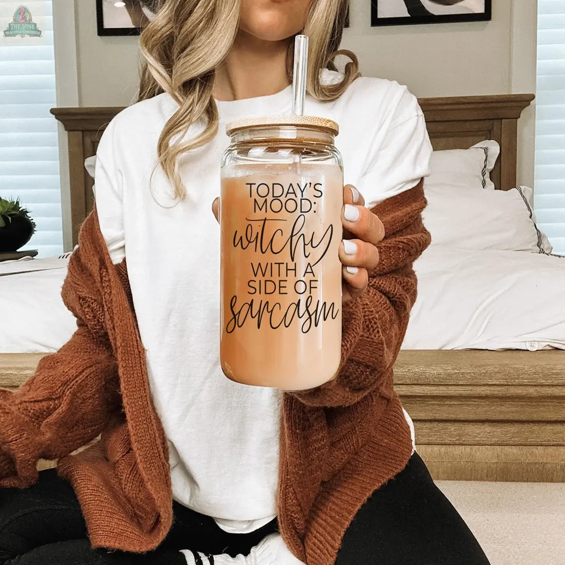A woman sits on a bed holding the Witchy 20oz glass tumbler with a straw. The jar reads, "Today's mood: witchy with a side of sarcasm." Her white shirt, brown cardigan, and wavy hair complete the cozy, witchy vibe.