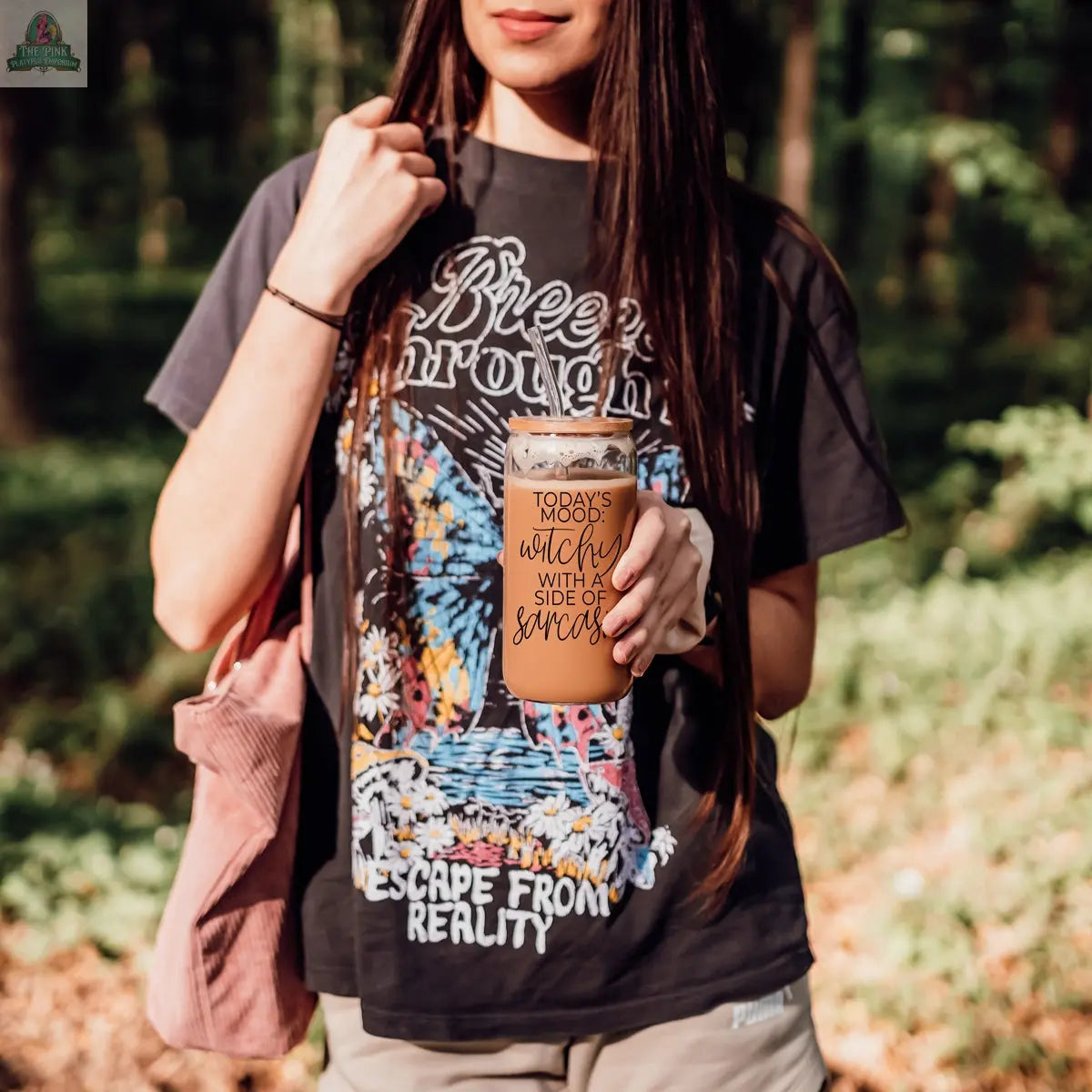 In a forest, a woman holds the Witchy 20oz eco-friendly tumbler that says, "Today’s mood: a touch of witch with a side of sarcasm." She sports a graphic tee and pink bag—a sassy gift for anyone with witchy vibes.