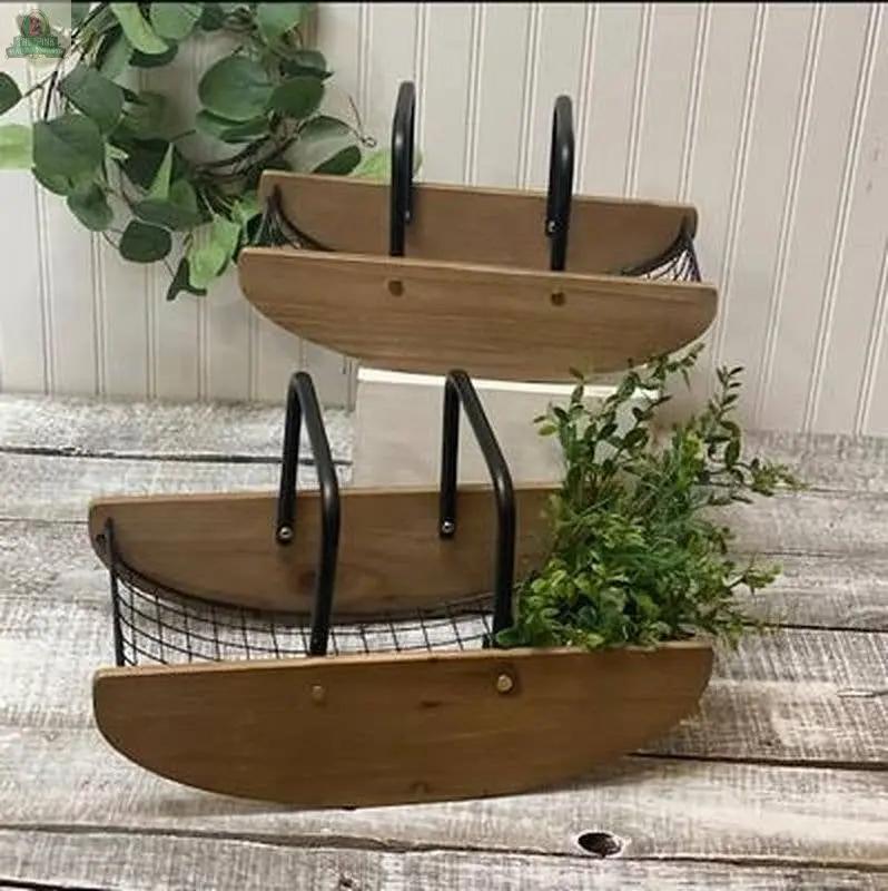 The Wood/Wire Rocking Baskets (Set of 2) feature half-moon shapes, black metal handles, and wire bottoms. Shown on a wooden surface with greenery, they are displayed against a white paneled wall.