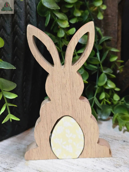 The Wooden Bunny with Enamel Egg Cutout 3D 5.5in features tall ears and a yellow floral enamel egg at its base, displayed on a light surface with green leafy plants for a charming spring decor accent.