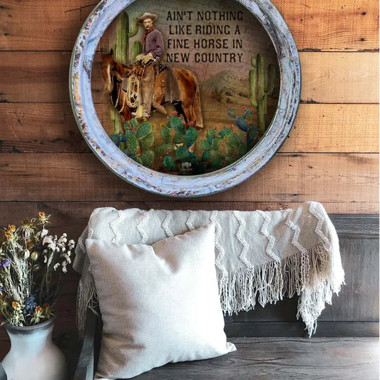 Decorative wall piece with cowboy scene and text above a rustic setting with pillows and a blanket.