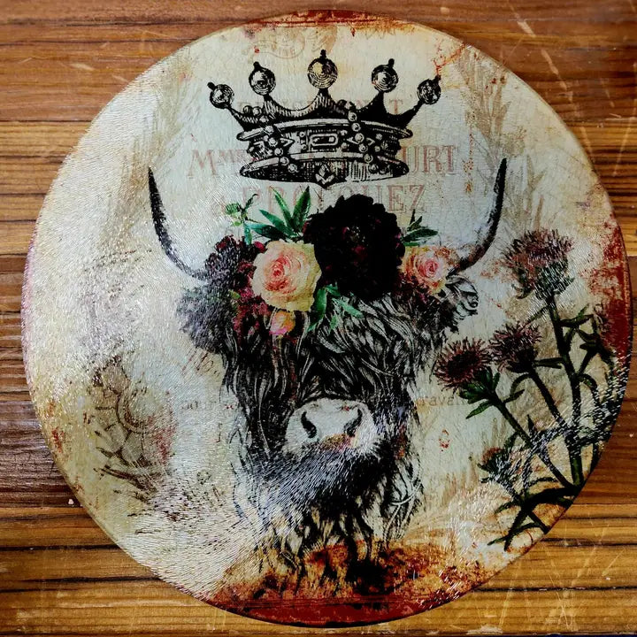 Round glass cutting board with a highland cow design, flowers, and a crown on a wooden surface.