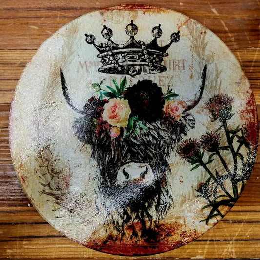 Round glass cutting board with a highland cow design, flowers, and a crown on a wooden surface.