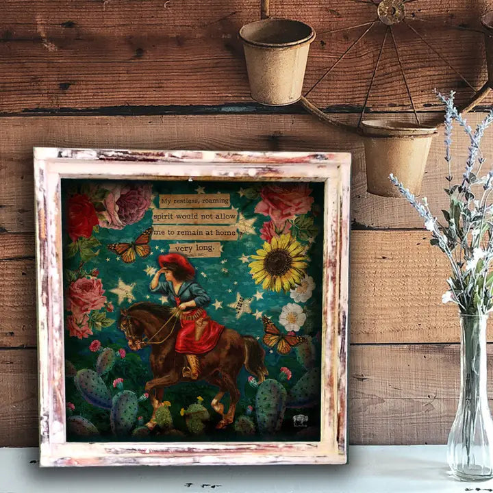 Framed artwork with a colorful scene of a person on horseback, flowers, and cacti against a wooden wall.