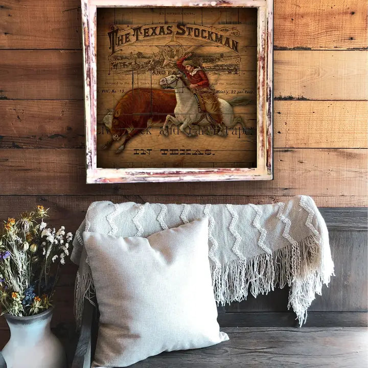 Framed vintage style advertisement for 'The Texas Stockman' on a wall with a wood background above a bench with a white blanket and pillow.