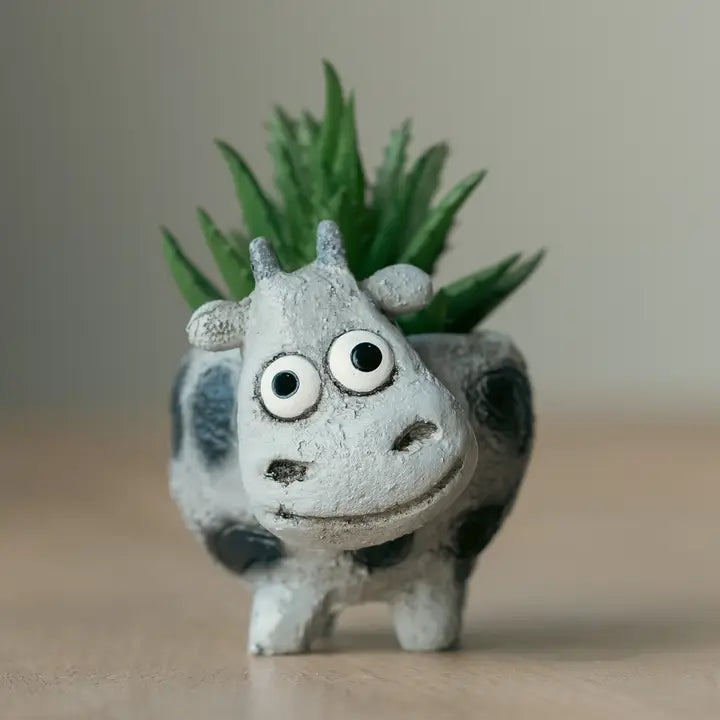 Ceramic cow succulent planter with large eyes and black spots, placed on a wooden surface with a blurred green succulent in the background.