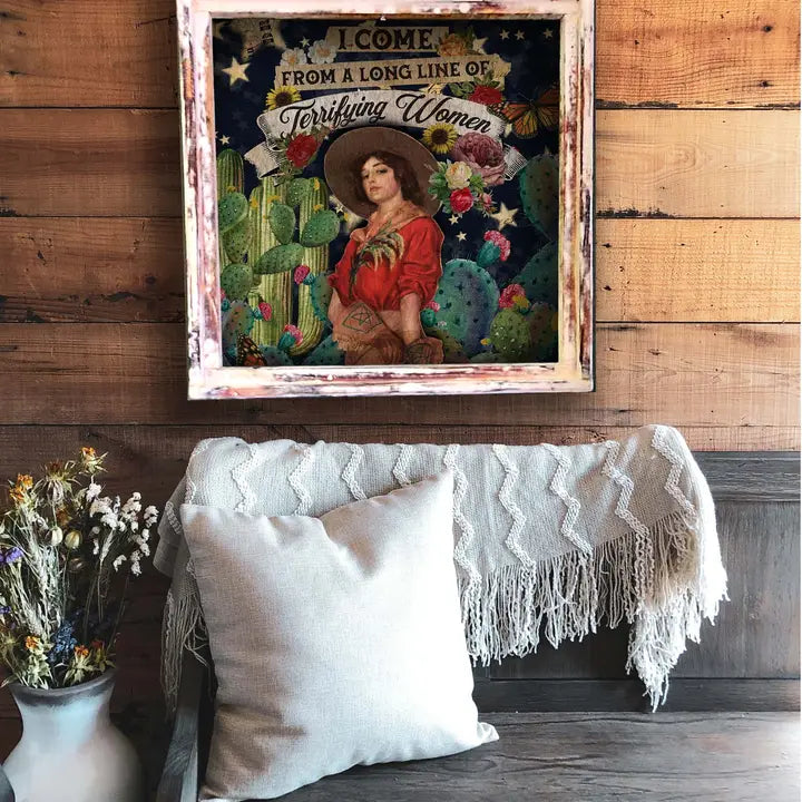 Decorative poster with a woman and cacti on a wooden wall above a couch with pillows and a blanket.