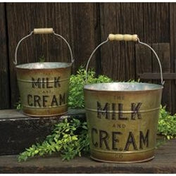 Two metal buckets labeled 'Milk and Cream' on a wooden surface with greenery.