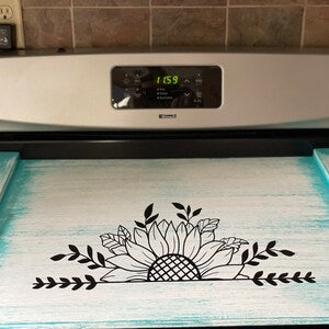 Pink Platypus Emporium rustic sunflower stove cover with black floral design on a distressed white solid wood farmhouse noodle board.